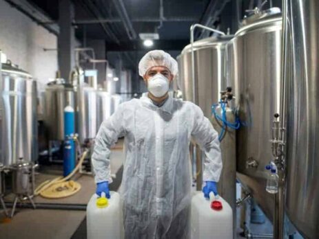 Worker wearing PPE carrying two plastic containers of chemicals standing next to chemical mixing tanks