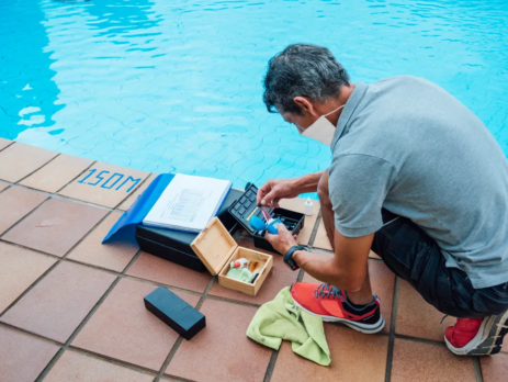 A man with a facemask on in a grey tshirt is checking the chlorine levels of a blue swimming pool while standing on tan-brown tiles besides it. Chlorine is an example of an anion.