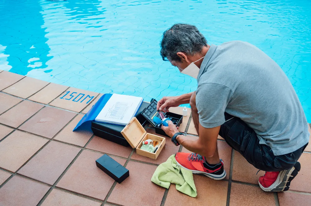 A man with a facemask on in a grey tshirt is checking the chlorine levels of a blue swimming pool while standing on tan-brown tiles besides it. Chlorine is an example of an anion.