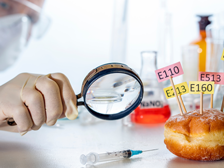 Scientist using a magnifying glass to look at a donut decorated with tablets with the names of E. food additives.