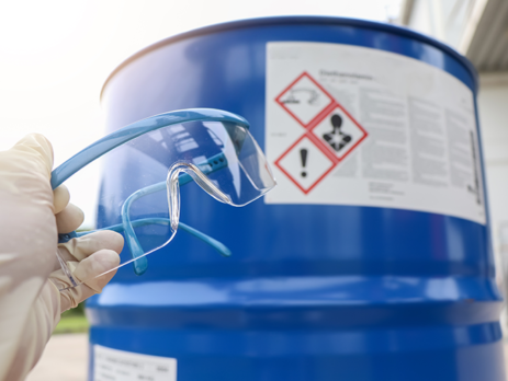 Person holding a pair of safety glasses next to a blue jerrycan of hazardous chemicals