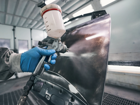 A blue-gloved hand spraying a chemical coating on a metal sheet.