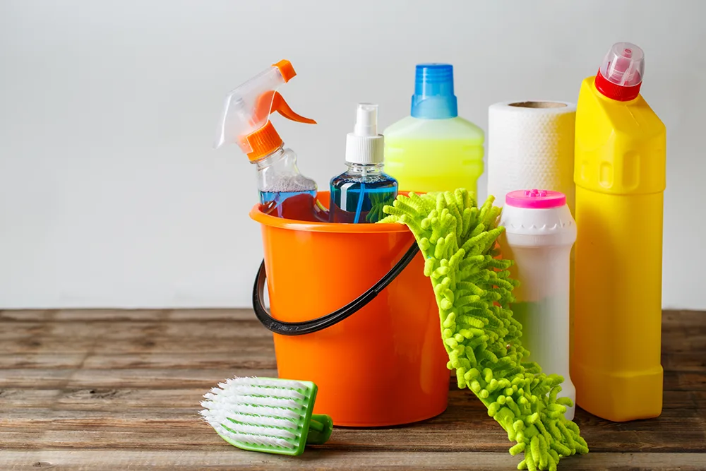 An orange bucket on a wooden table filled with various cleaning chemicals, including bleach, ammonia, and ethanol.