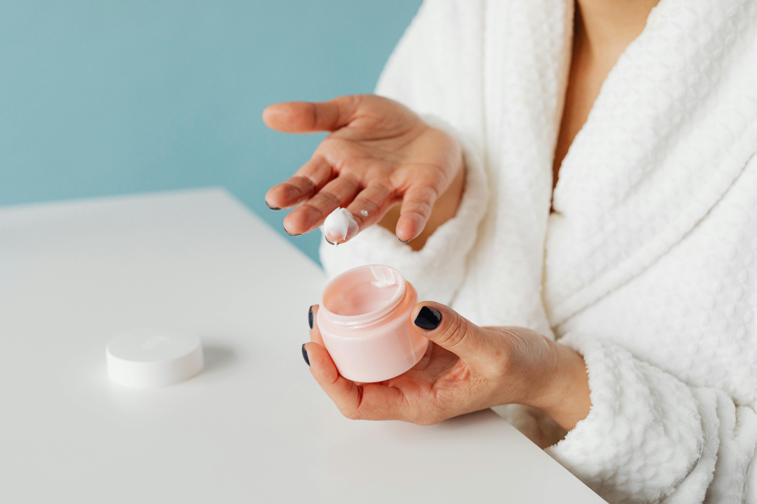 A person in a white dressing gown applies anti-ageing cream to their finger. They are sitting at a white table with a blue backdrop.