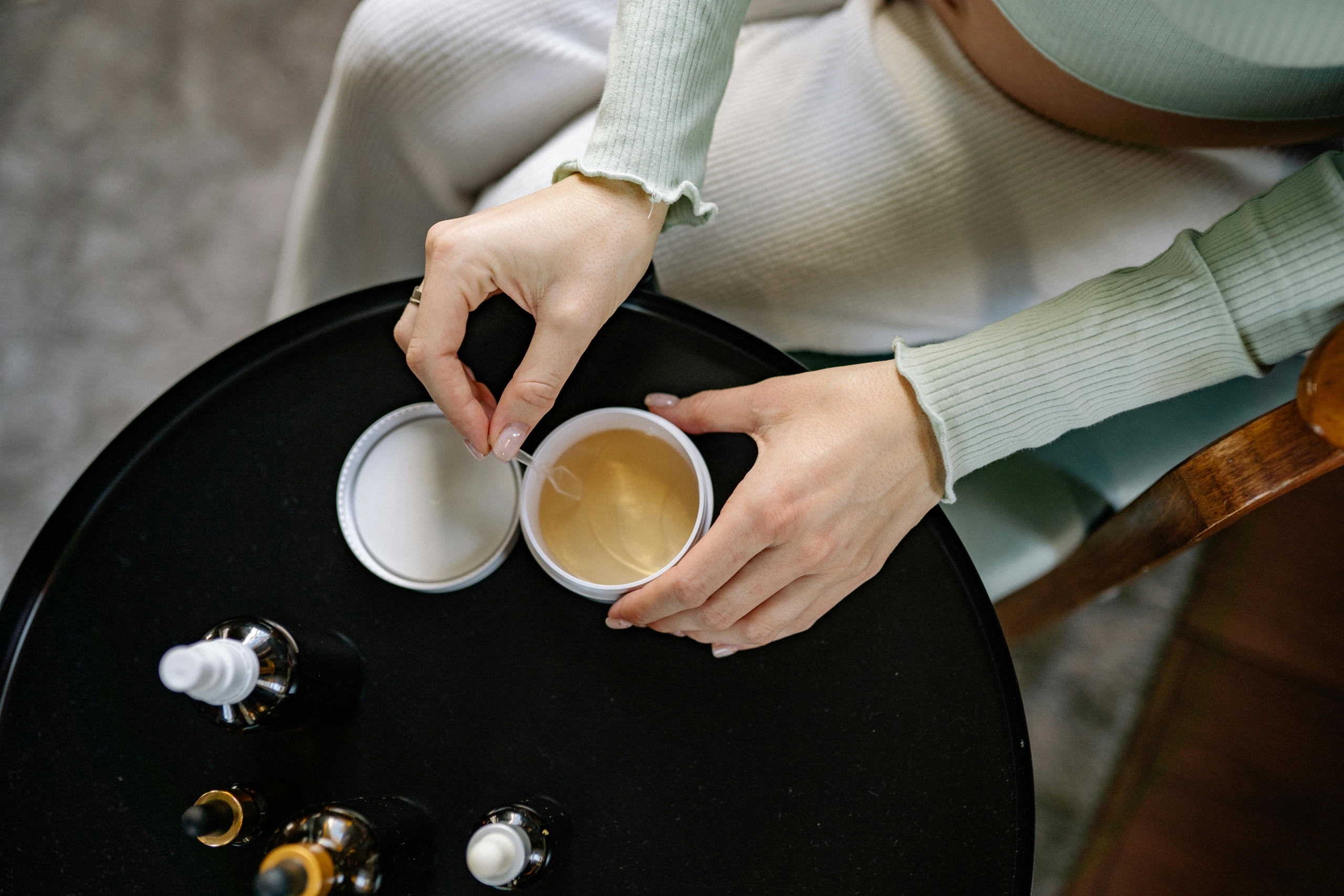 A person sits at a table, preparing anti-ageing cosmetics with a spatula. In their hand is a small tub of one product, while on the black table lies others in glass vials.