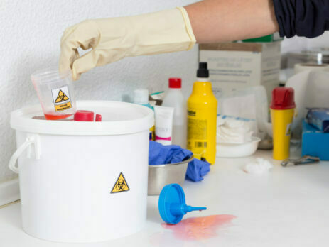 Scientist wearing gloves throwing away lab waste into a small plastic hazardous waste bucket.||Scientist wearing blue gloves carrying a red plastic bag with a public biohazard sign.||Laboratory waste in a bin.
