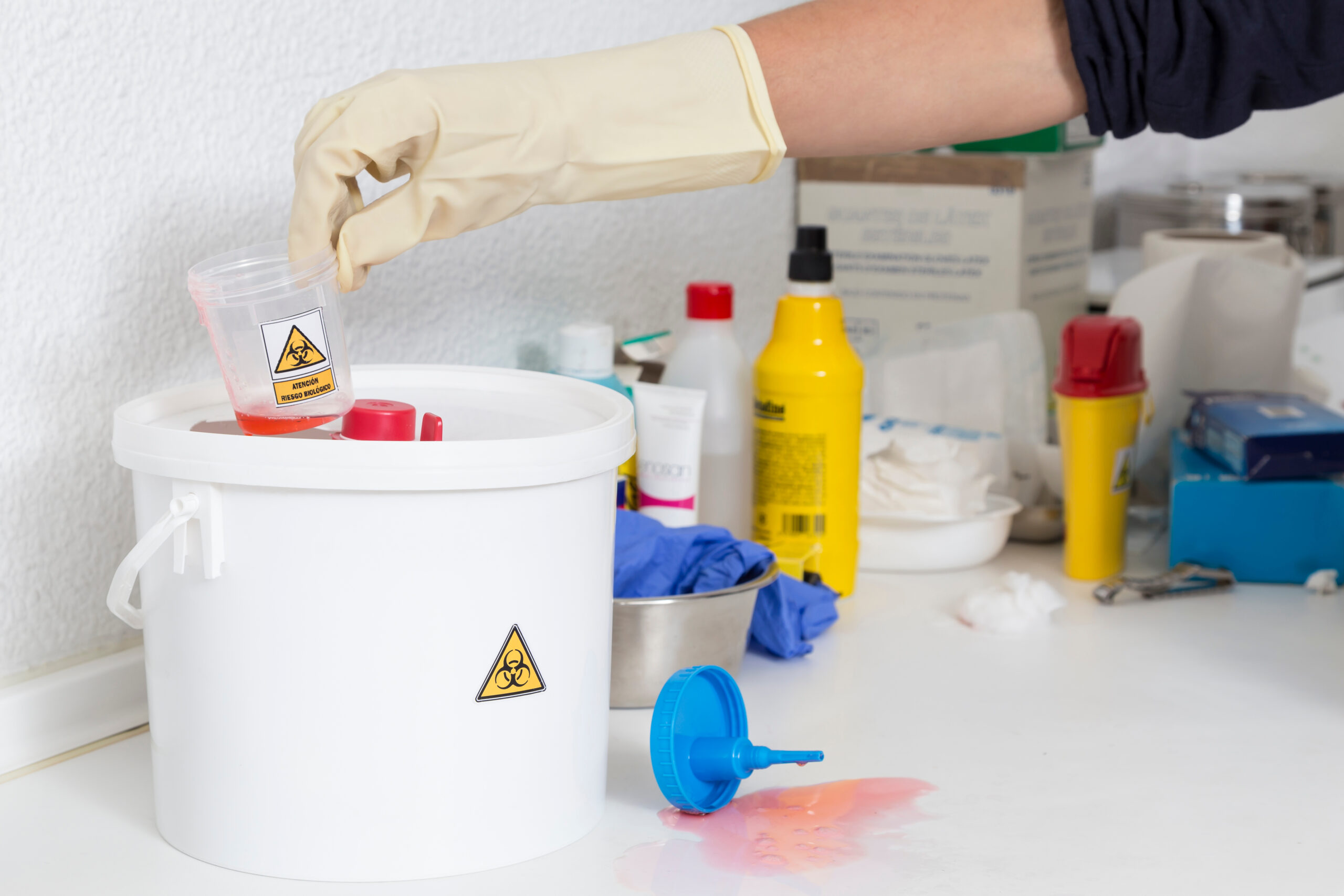Scientist wearing gloves throwing away lab waste into a small plastic hazardous waste bucket.||Scientist wearing blue gloves carrying a red plastic bag with a public biohazard sign.||Laboratory waste in a bin.