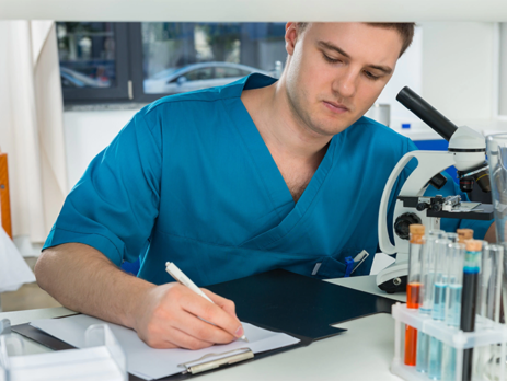 Young male student in scrubs writing research notes after looking through a microscope in a lab||The steps to getting into medical school||Female doctor wearing a mask checking on an elderly female patient||A male GP taking an older man's blood pressure||The steps to getting into medical school