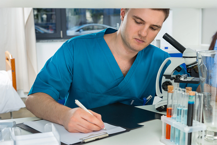 Young male student in scrubs writing research notes after looking through a microscope in a lab||The steps to getting into medical school||Female doctor wearing a mask checking on an elderly female patient||A male GP taking an older man's blood pressure||The steps to getting into medical school